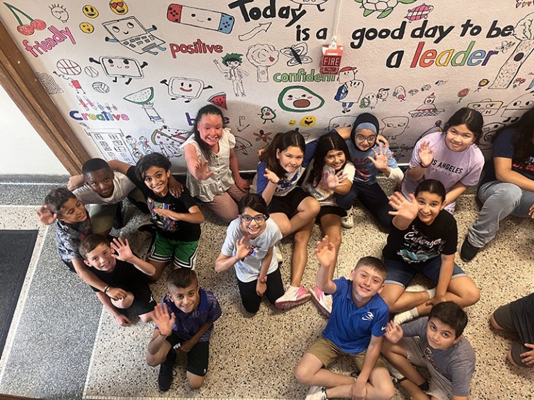 A group of students poses in front of a mural they are creating as part of the 2024 Summer Discovery Program at Lindbergh Elementary.
